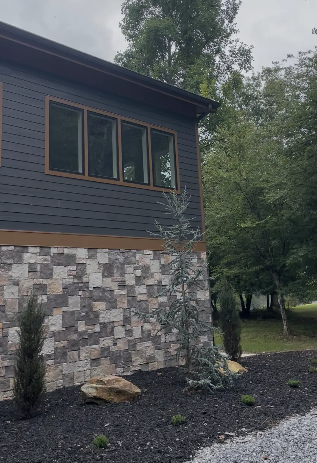 Side of a new house with a stone base, dark siding and fresh landscaping